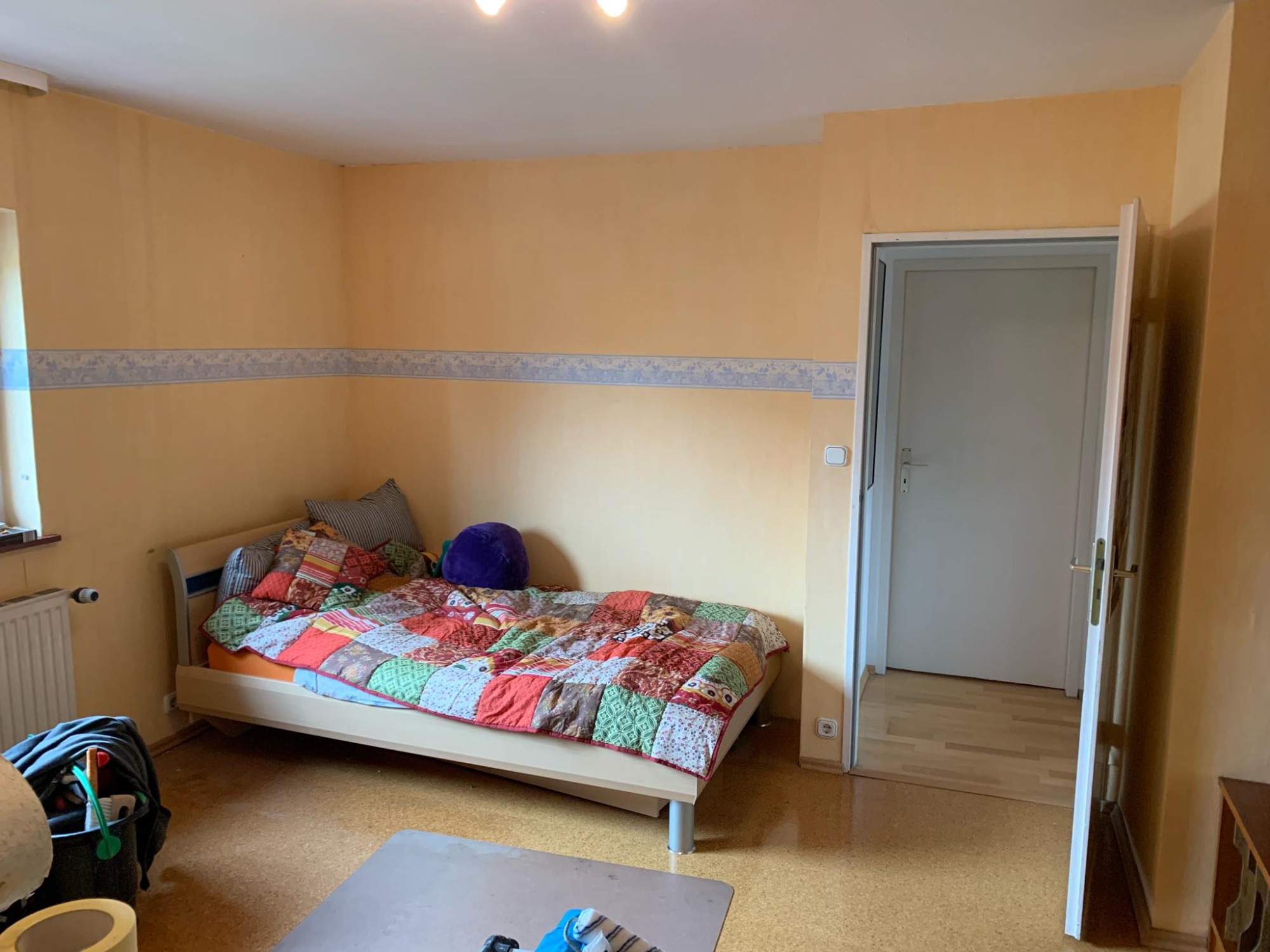 Pokój z żółtymi ścianami, ozdobną bordiurą, łóżkiem z patchworkową narzutą i otwartymi drzwiami do innego pomieszczenia z parkietem.