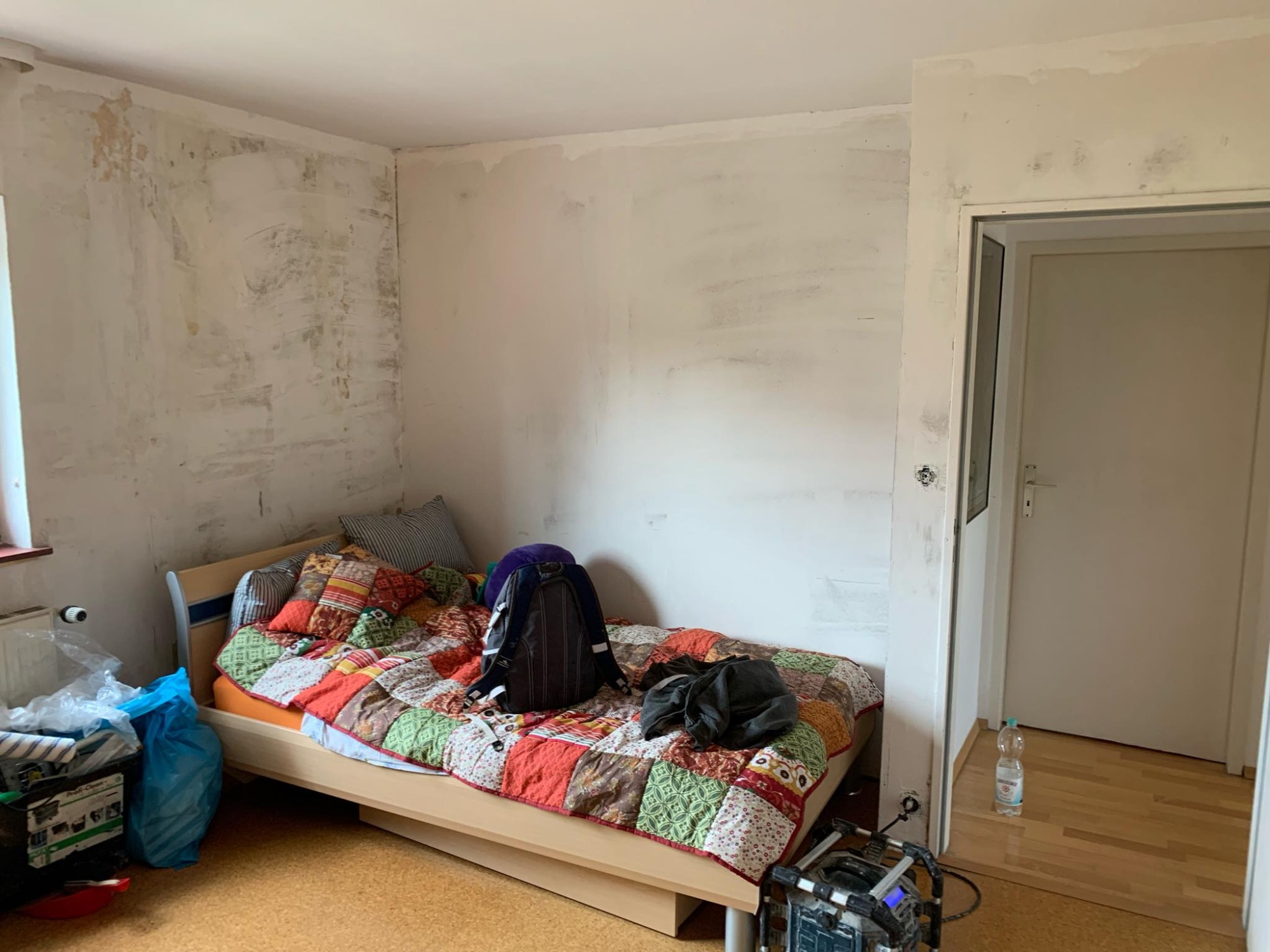 Pokój w trakcie przygotowania do malowania: ściany po szpachlowaniu, widoczne ślady po wcześniejszych warstwach, zakryte meble, na podłodze butelka wody i sprzęt budowlany.