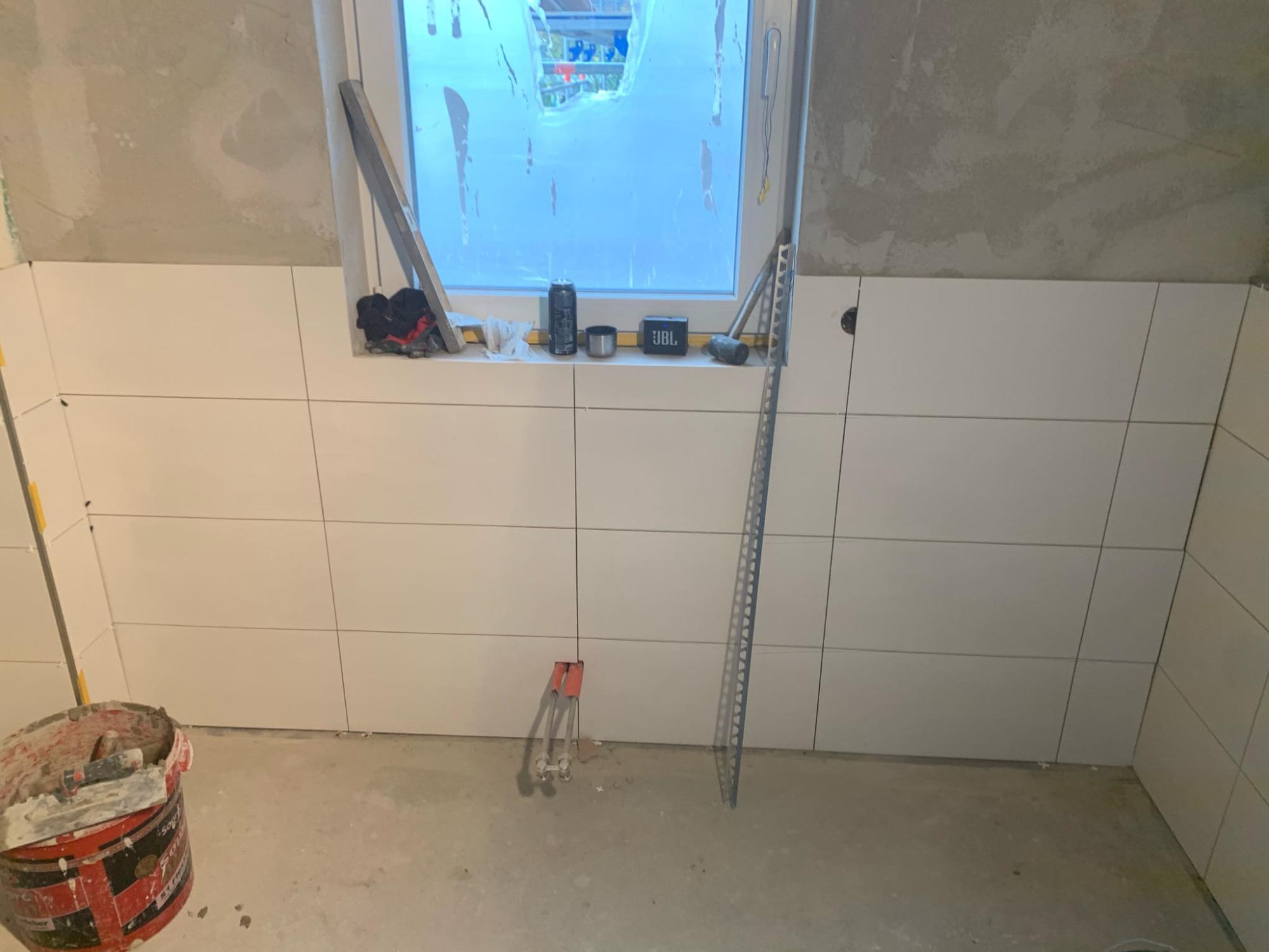 Ściana łazienki w trakcie układania białych, prostokątnych płytek, widoczne narzędzia glazurnicze, poziomica, puszka napoju, głośnik JBL na parapecie okna, surowy beton na ścianie powyżej płytek.