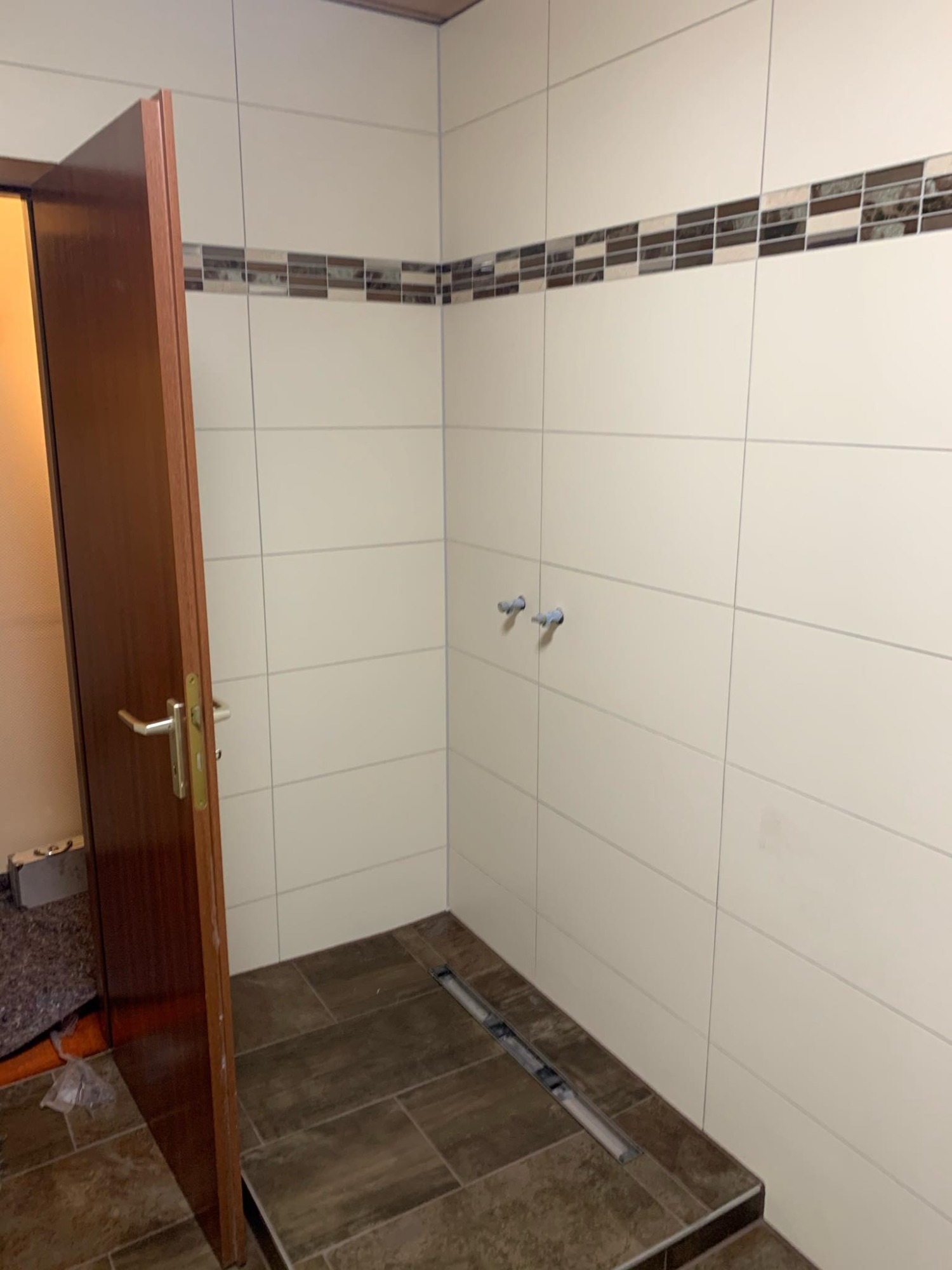 Nowa łazienka w trakcie wykańczania: białe płytki ścienne z dekoracyjnym pasem mozaiki, brązowe płytki podłogowe z odpływem liniowym, otwarte drewniane drzwi.