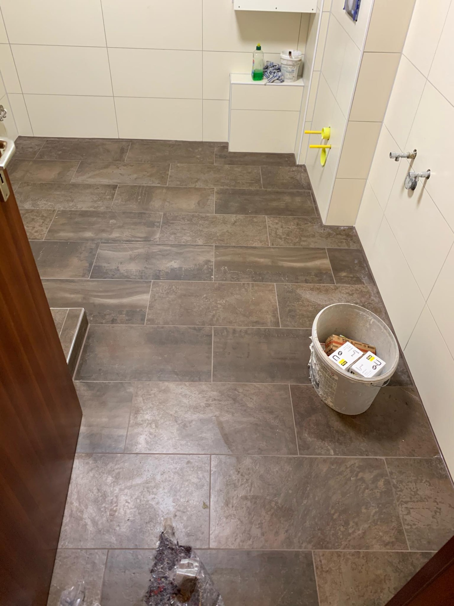 Ułożona podłoga z płytek imitujących drewno w łazience, widoczny otwarty brązowy drzwi, białe płytki na ścianach, wiadro z opakowaniami po kleju i resztki po pracy na podłodze.