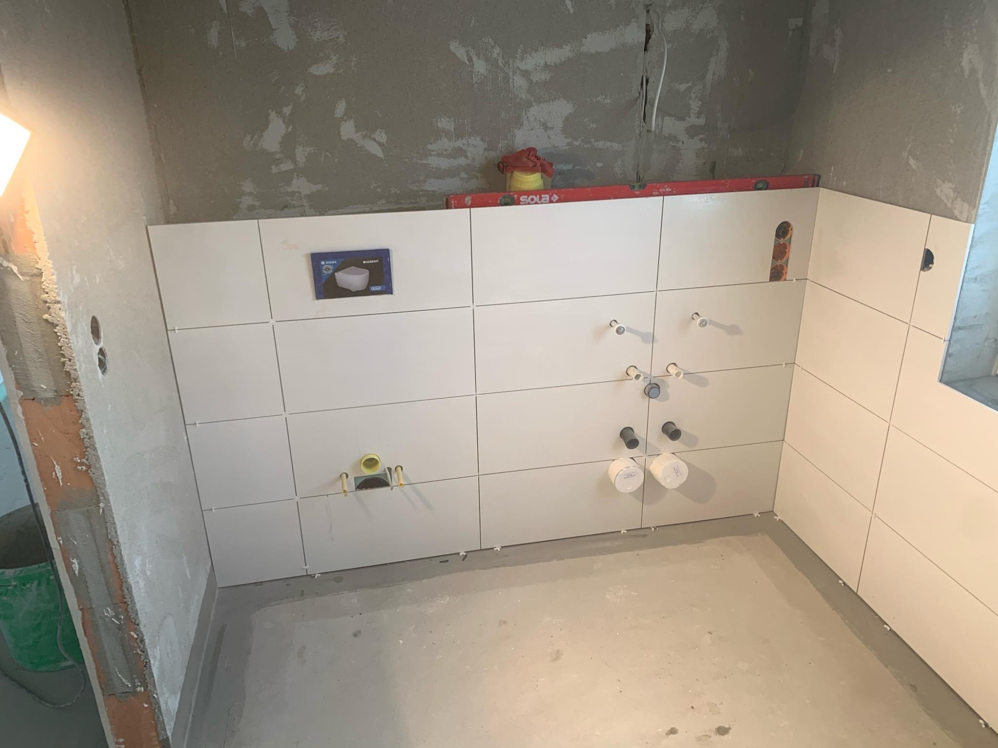 Ściana w łazience w trakcie układania białych, prostokątnych płytek; widoczne wystające rury instalacyjne i poziomica Sola na górze ściany.