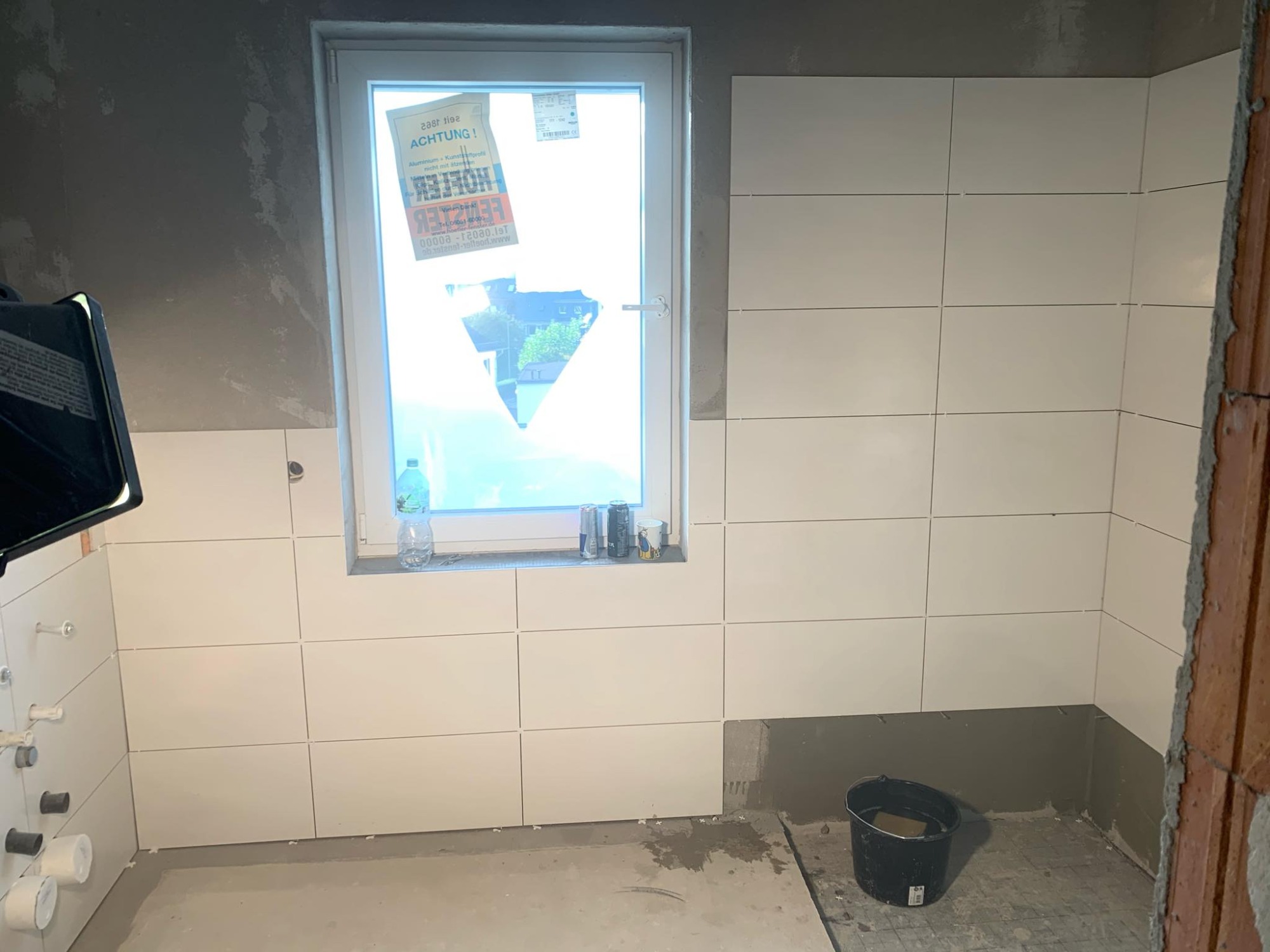 Białe, prostokątne płytki ułożone na ścianie w łazience w Lublinie, widoczne niedokończone prace, surowe ściany i podłoga, okno z naklejkami, wiadro na podłodze.