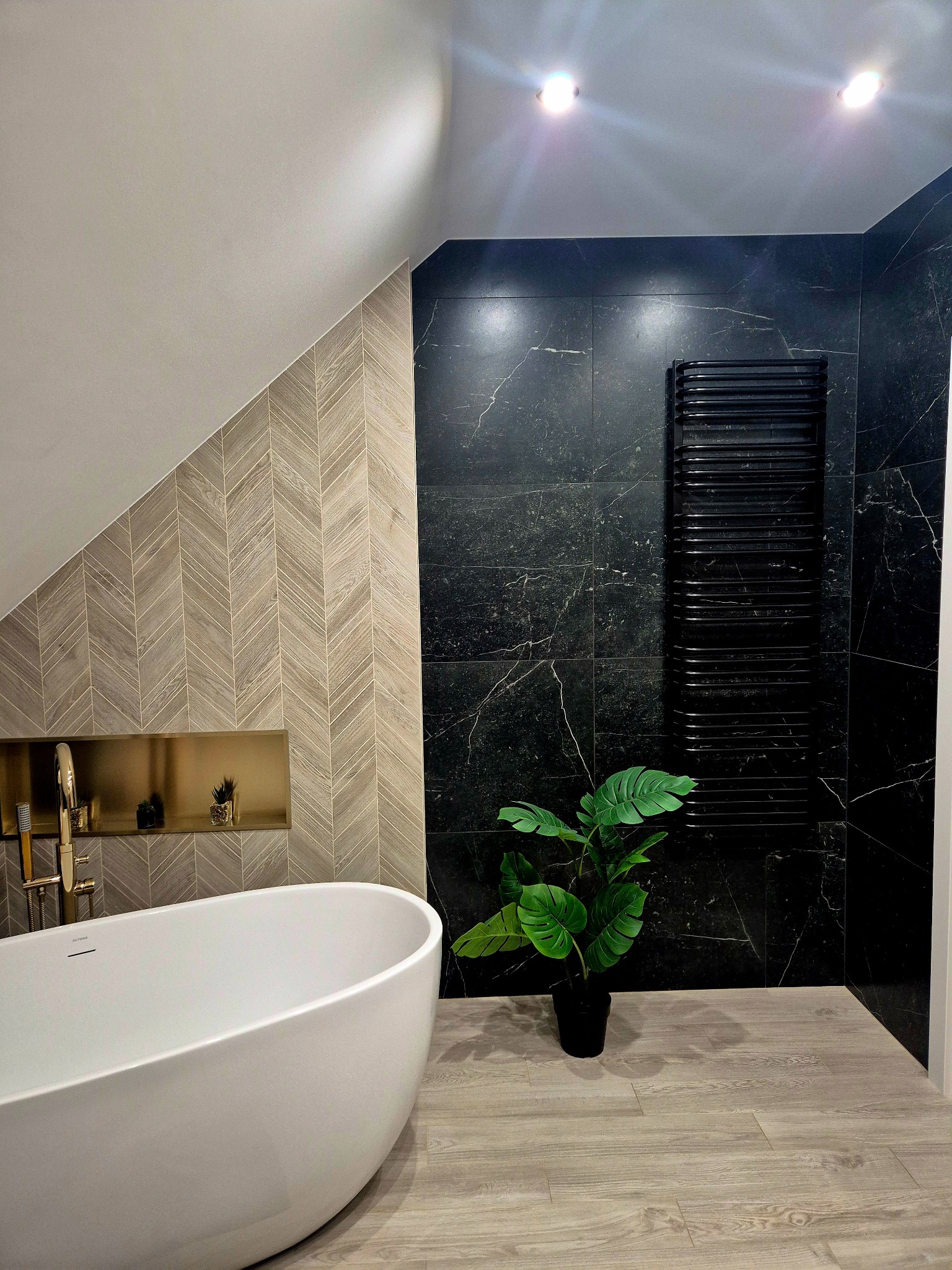 Nowoczesna łazienka na poddaszu z wolnostojącą wanną, czarnymi płytkami imitującymi marmur i grzejnikiem drabinkowym. Uwagę przykuwają płytki ułożone w jodełkę.