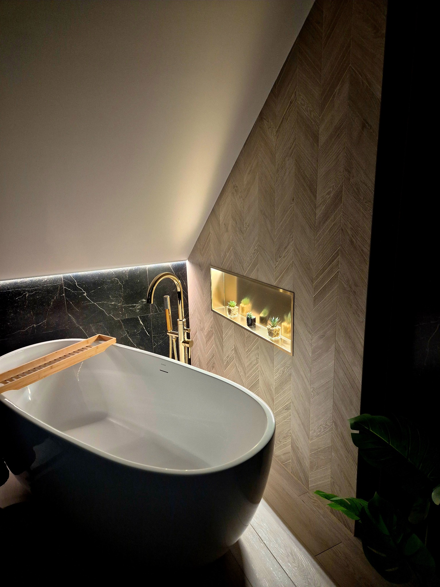Nowoczesna łazienka na poddaszu: wolnostojąca wanna, złota armatura, marmurowe płytki i drewnopodobna ściana w jodełkę z podświetlaną wnęką na dekoracje.