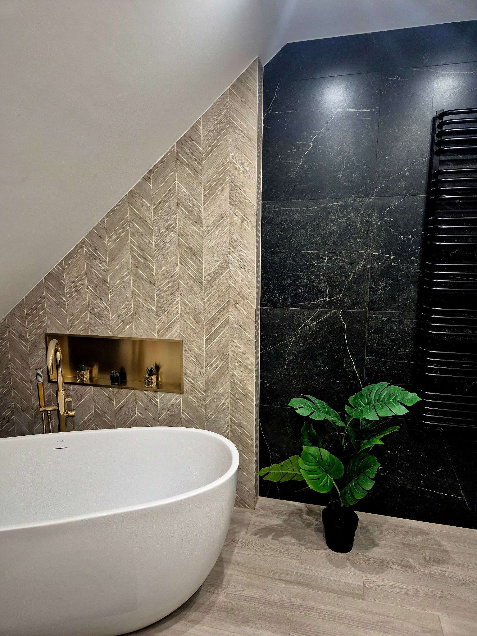 Nowoczesna łazienka na poddaszu: biała wanna wolnostojąca, płytki w jodełkę, czarne marmurowe ściany i dekoracyjna roślina. Stylowe wykończenie.