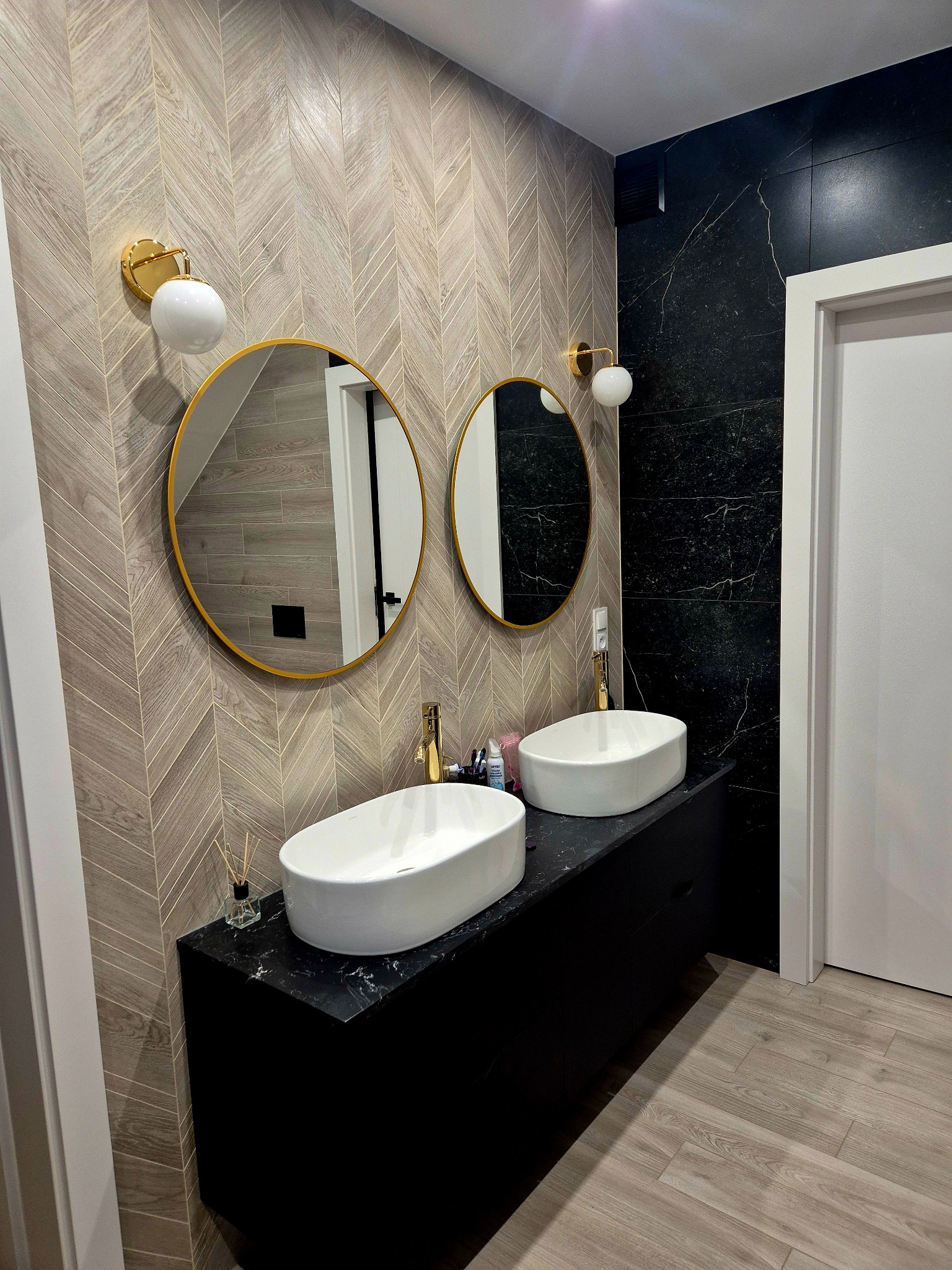 Stylowa łazienka z dwiema owalnymi umywalkami na czarnym blacie, okrągłymi lustrami w złotych ramach i wzorzystą, drewnopodobną okładziną ścienną.