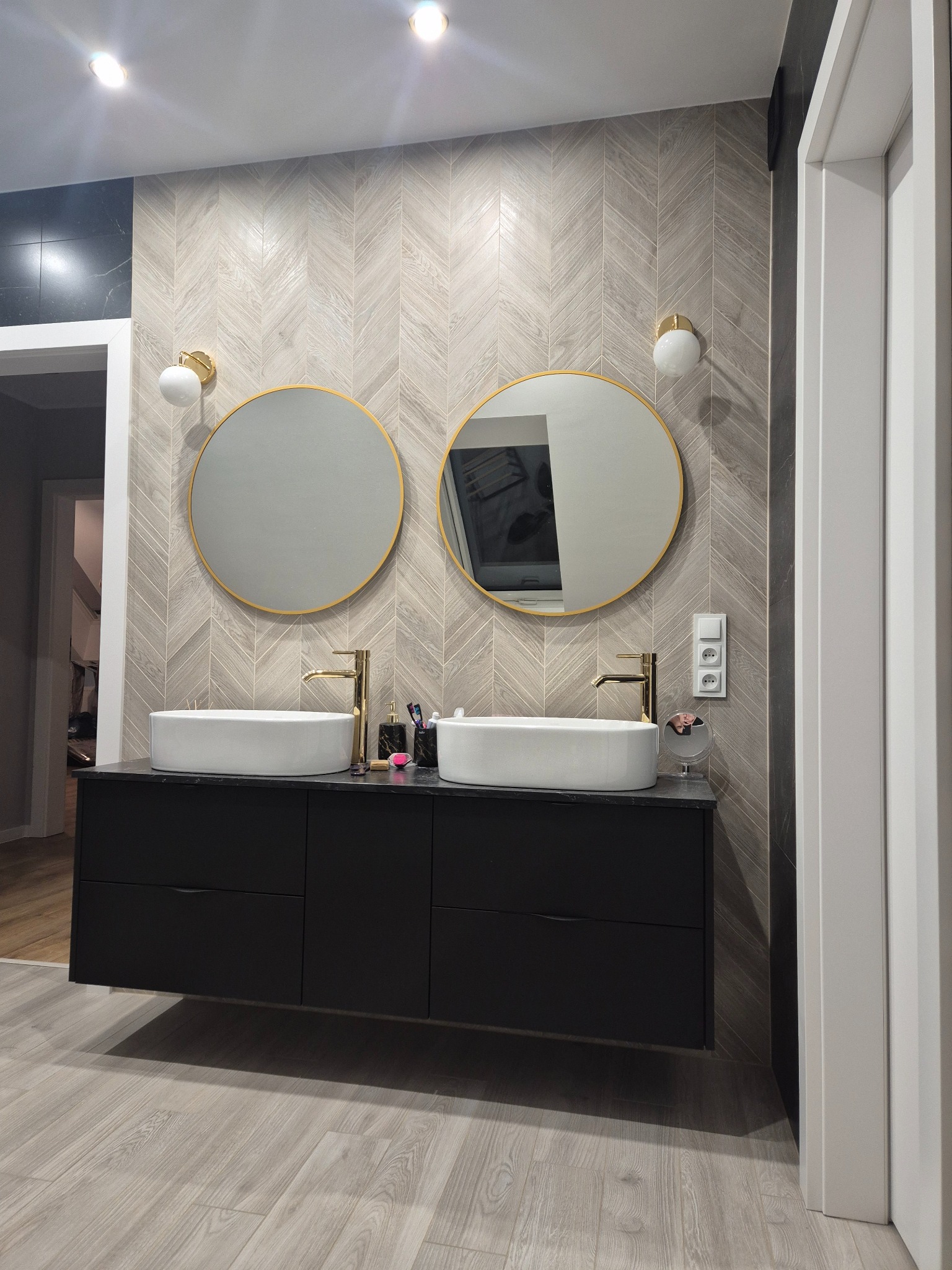 Nowoczesna łazienka z dwoma okrągłymi lustrami w złotych ramach nad czarną szafką z umywalkami. Ściana wyłożona płytkami w jodełkę, podłoga imitująca drewno.