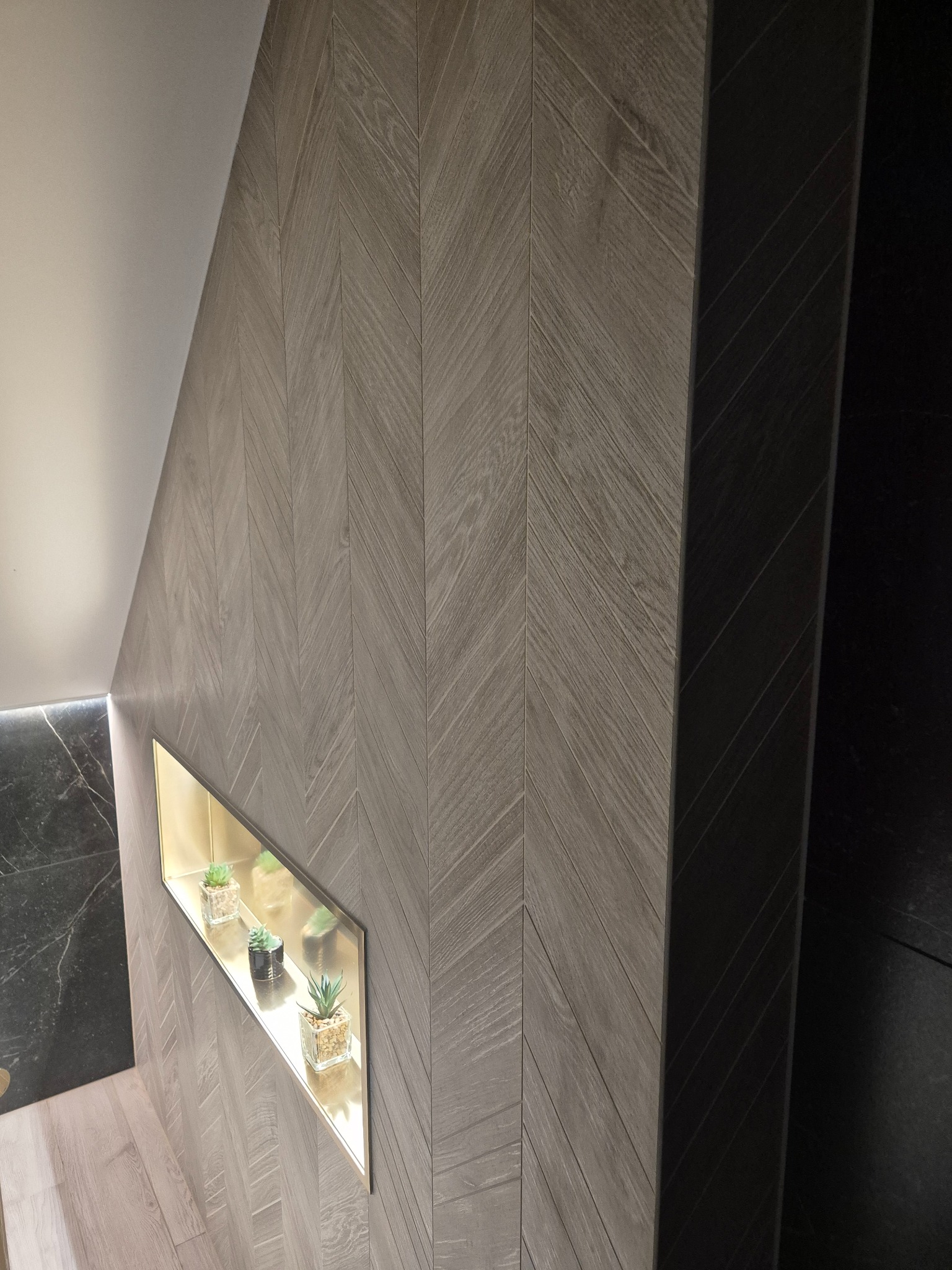 Ściana wyłożona płytkami w jodełkę, z wnęką oświetloną złotym światłem, w której umieszczono małe rośliny doniczkowe. Minimalistyczny design łazienki.