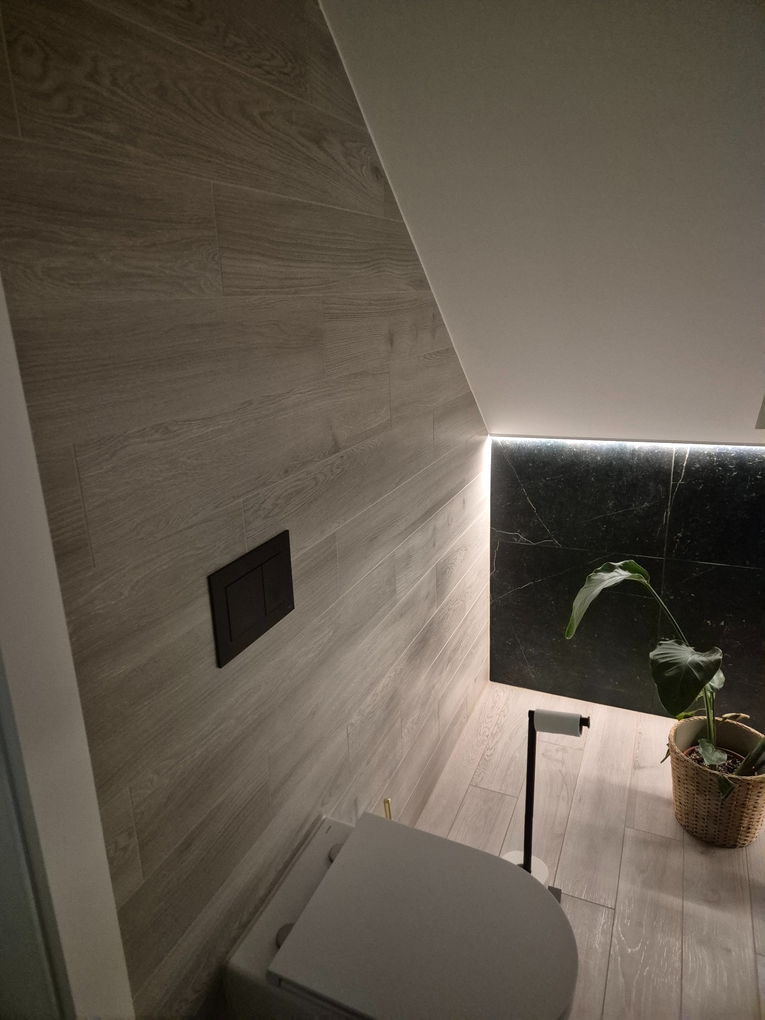 Nowoczesna łazienka z drewnopodobnymi płytkami na ścianach i podłodze, czarnym marmurem podświetlonym LED, minimalistyczną armaturą i rośliną w koszu.
