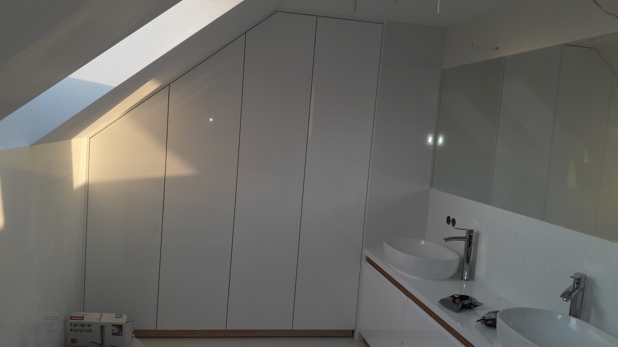 Biała łazienka na poddaszu ze skośnym sufitem, zabudowa szaf wnękowych z połyskiem, dwie owalne umywalki nablatowe z chromowanymi bateriami.