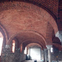 Kopuła z cegły klinkierowej w zabytkowym budynku, wsparta na kolumnach, z widocznymi oknami i fragmentem kamiennej ściany, w tle brama garażowa.