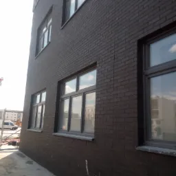 Ciemnobrązowa elewacja budynku z cegły klinkierowej, widoczne okna z szarymi ramami i fragment chodnika w trakcie budowy.