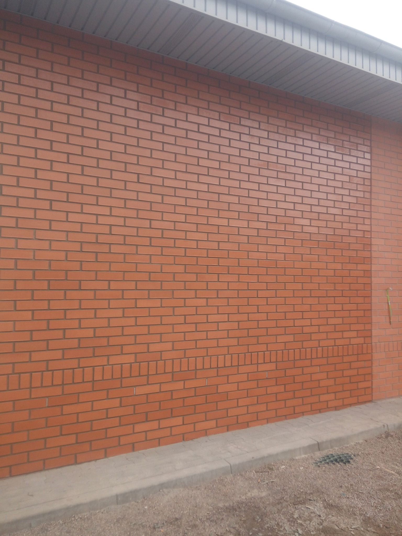 Świeżo wymurowana ściana z cegły klinkierowej w Kowala-Stępocinie, widoczny fragment dachu i chodnik z kostki brukowej.