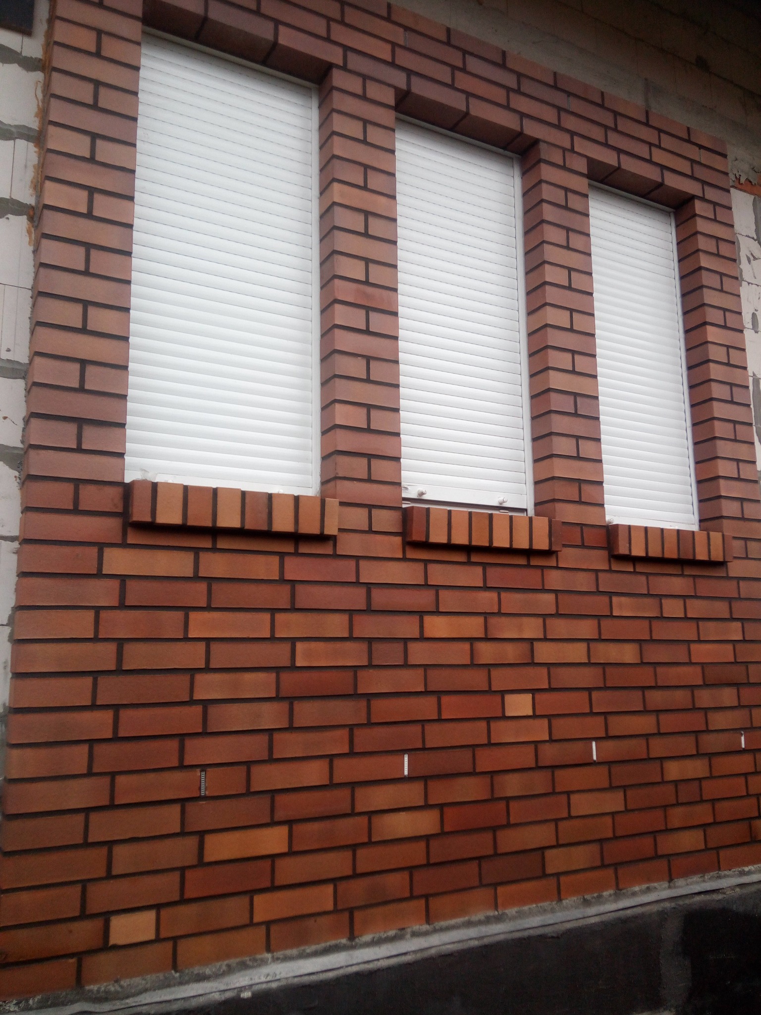 Elewacja budynku z czerwonej cegły klinkierowej z trzema oknami z żaluzjami zewnętrznymi, widoczne fragmenty ocieplenia budynku.