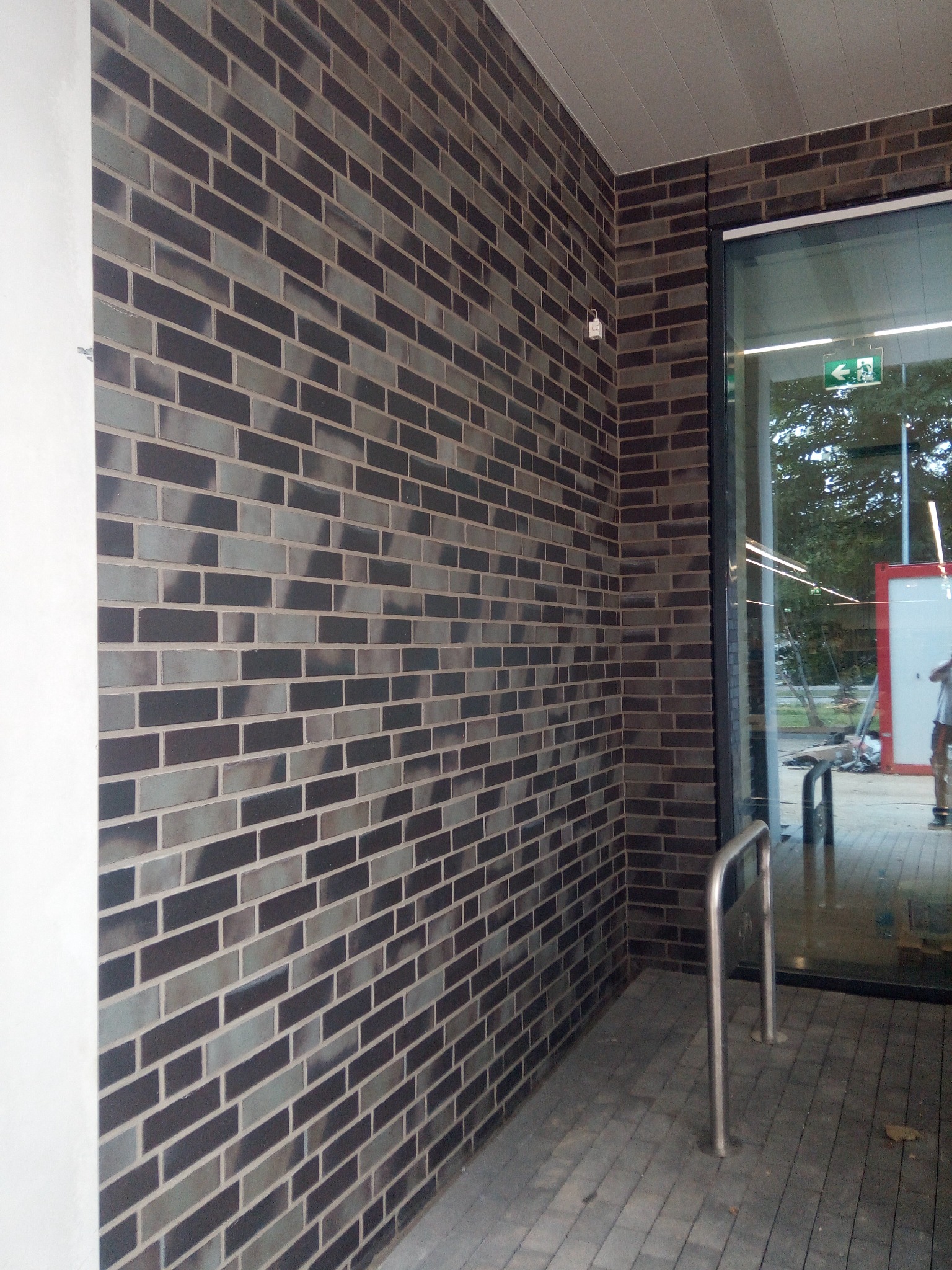 Elewacja z ciemnej, cieniowanej cegły klinkierowej przy wejściu do budynku z widocznym fragmentem chodnika z kostki brukowej i elementem metalowej barierki