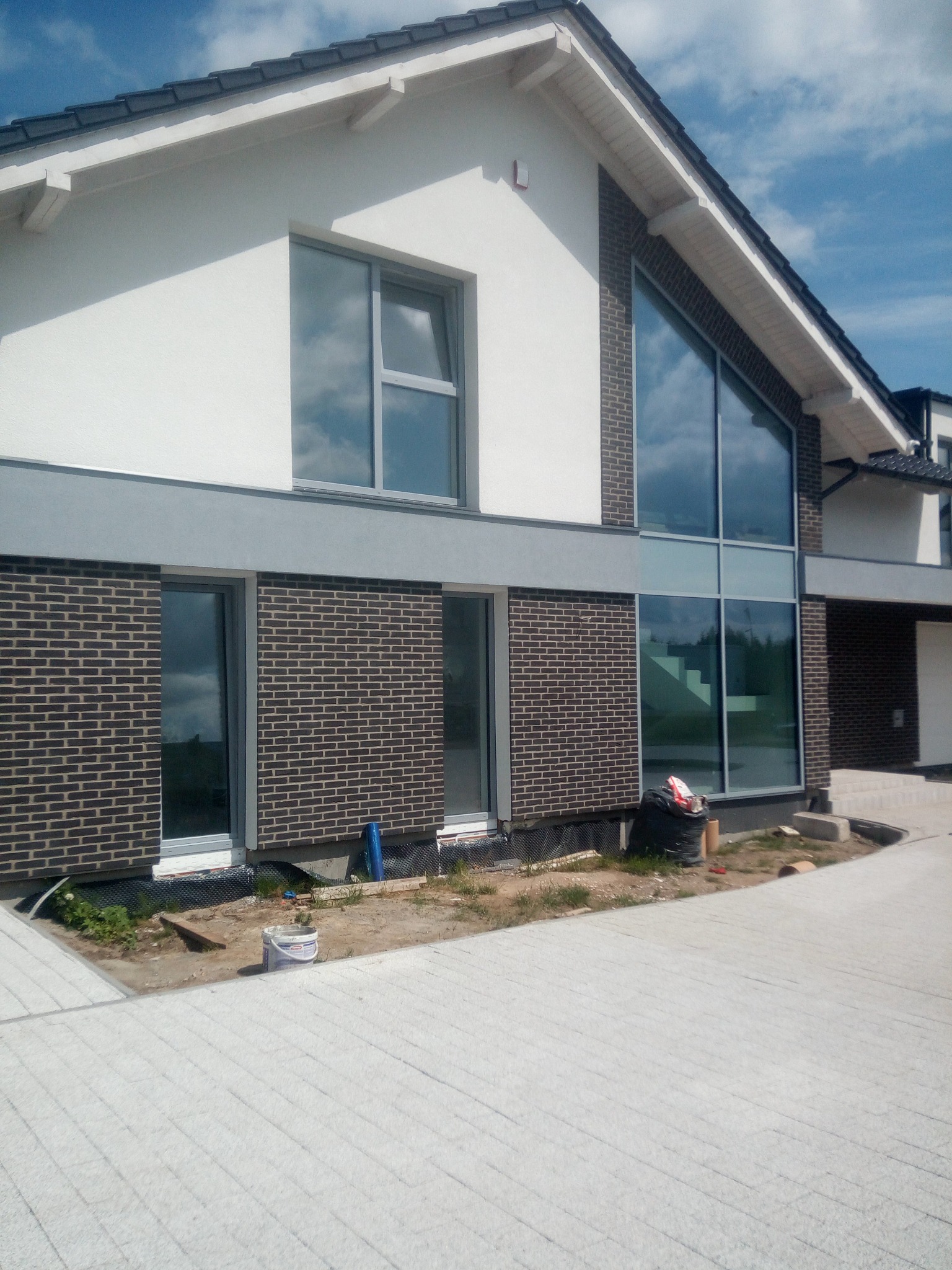 Nowoczesny dom z elewacją z cegły klinkierowej i dużymi oknami, fragment chodnika z kostki brukowej, widoczne prace wykończeniowe.
