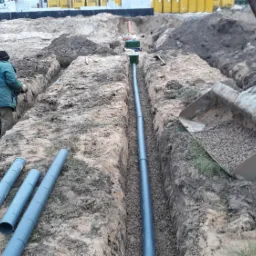 Wykopany rów z ułożonymi rurami drenażowymi, pracownik w zielonej kurtce w rowie, mini koparka z łyżką wypełnioną żwirem w tle, przygotowane rury na ziemi, teren budowy.