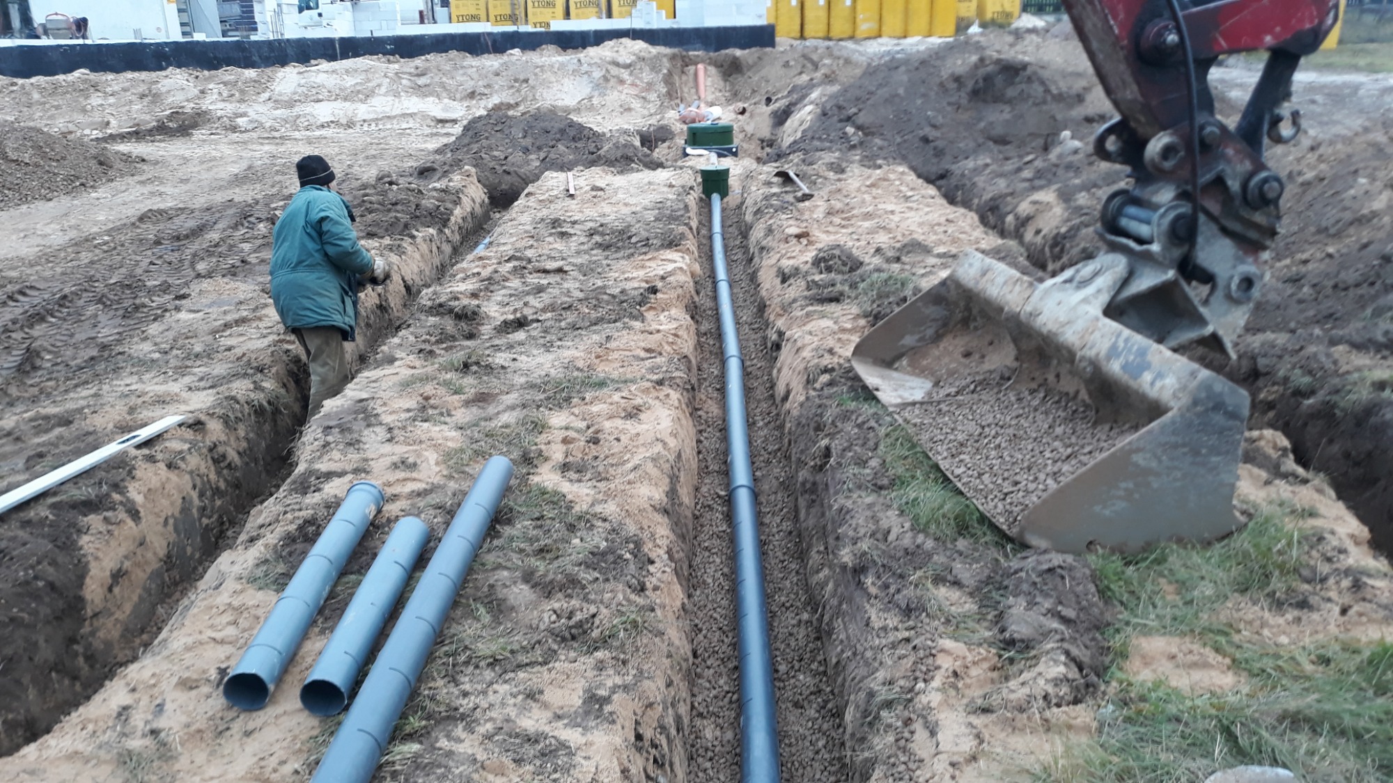 Wykopany rów z ułożonymi rurami drenażowymi, pracownik w zielonej kurtce w rowie, mini koparka z łyżką wypełnioną żwirem w tle, przygotowane rury na ziemi, teren budowy.