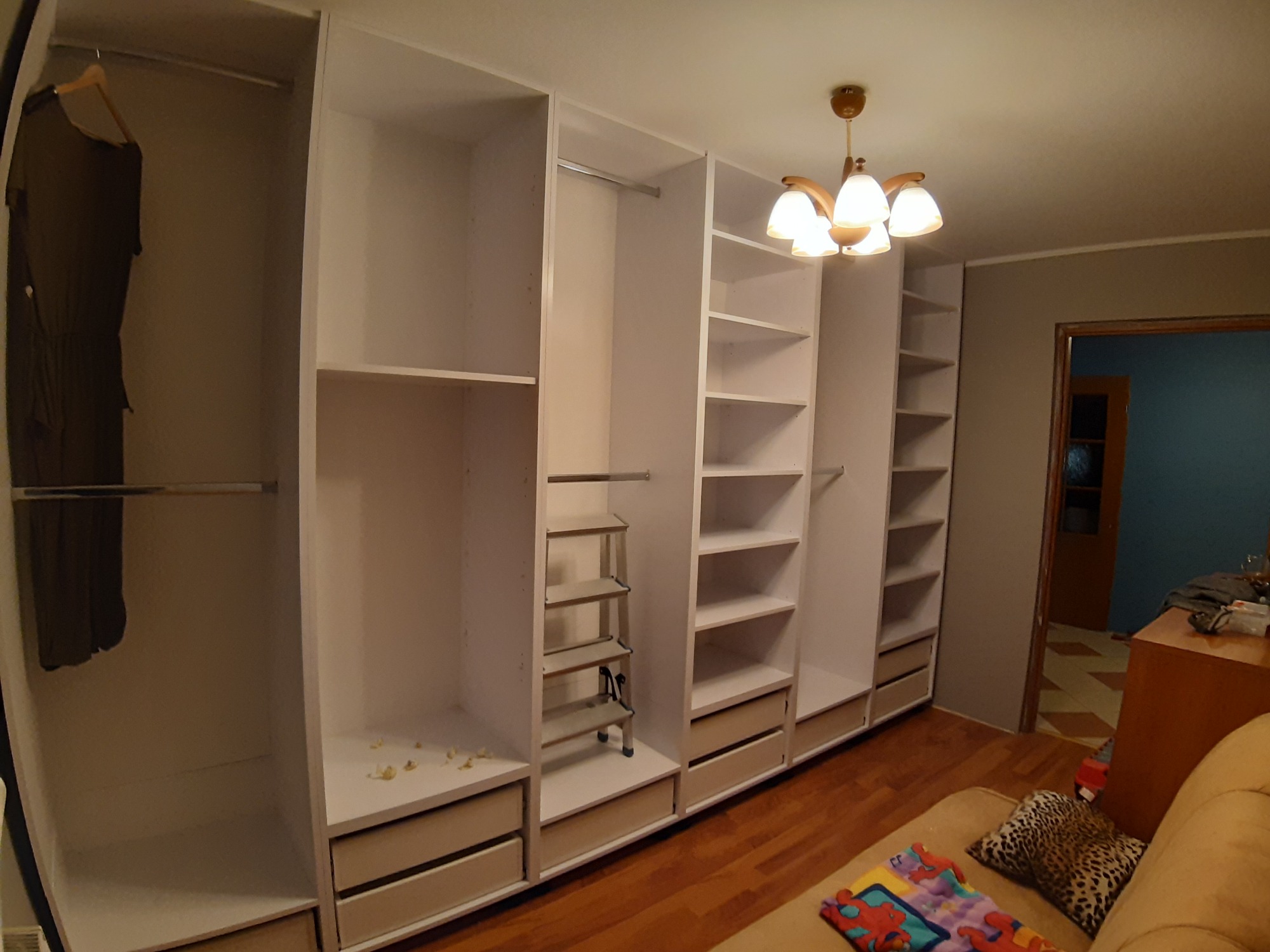 Biała, modułowa garderoba z półkami, szufladami i drążkami na ubrania, oświetlona ciepłym światłem lampy sufitowej, z widoczną odzieżą wiszącą na wieszaku.