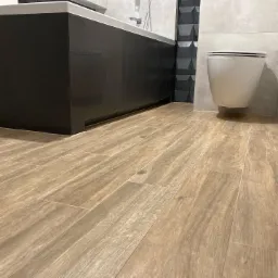 Nowoczesna łazienka z drewnopodobną podłogą z płytek, podwieszaną toaletą i czarną zabudową wanny.