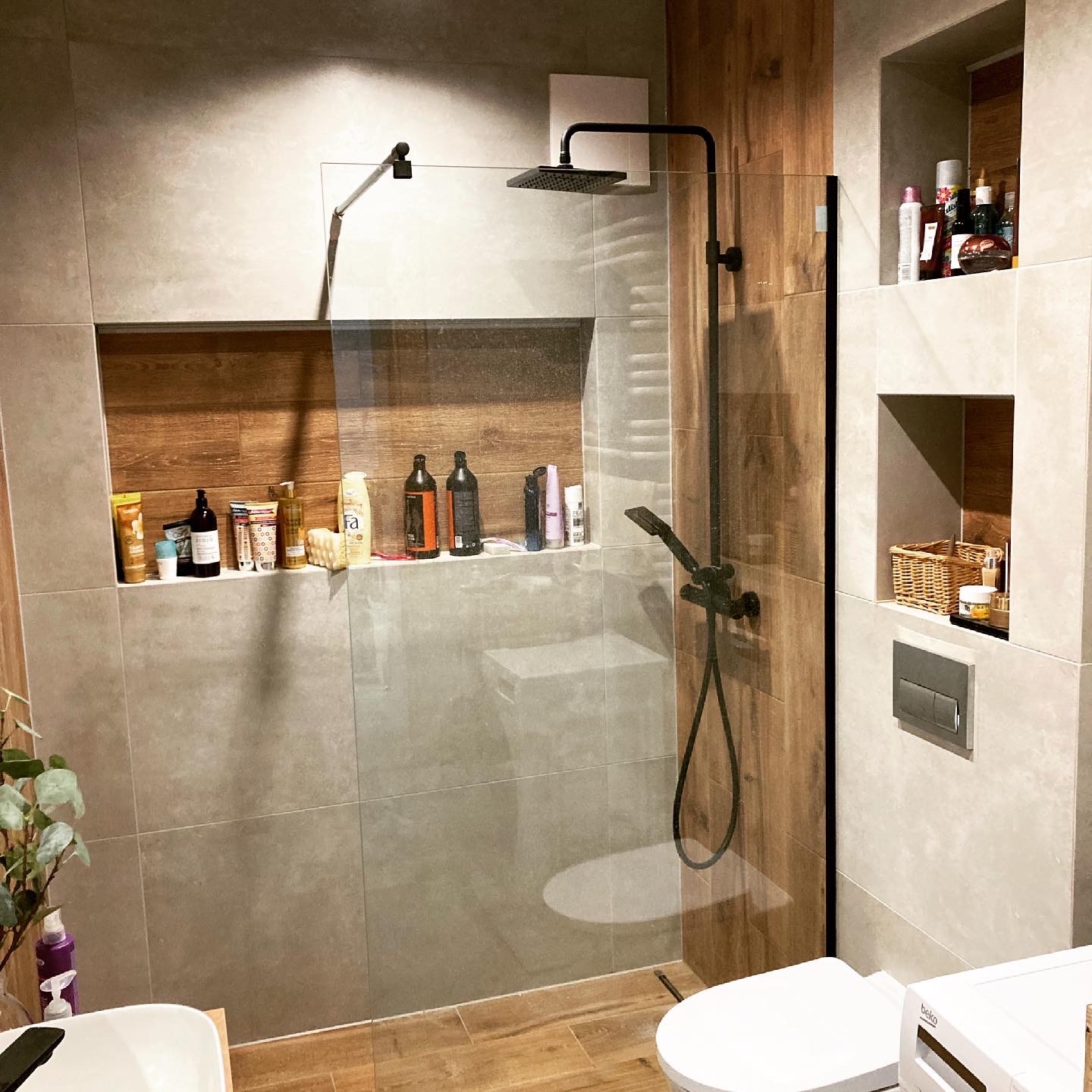 Nowoczesna łazienka z kabiną prysznicową typu walk-in, wykończona płytkami w odcieniach szarości i drewna, z czarną armaturą i wnękami na kosmetyki.
