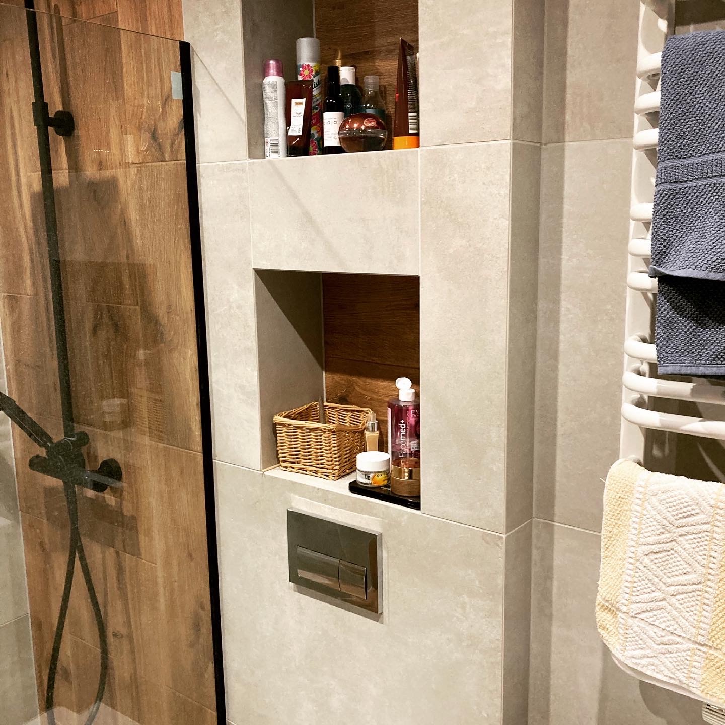 Nowoczesna łazienka z kabiną prysznicową z drewnopodobnymi płytkami, wnękami w ścianie wyłożonymi kosmetykami i koszykiem, oraz grzejnikiem drabinkowym z ręcznikami.