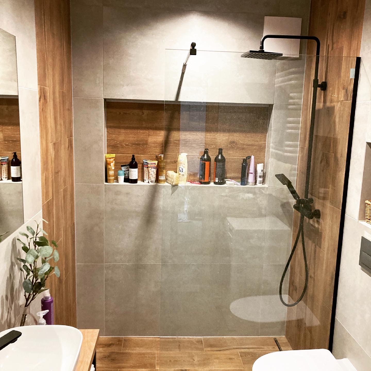 Nowoczesna łazienka z kabiną prysznicową typu walk-in, wykończona płytkami w odcieniach szarości i drewna, z wnęką na kosmetyki i czarną armaturą.