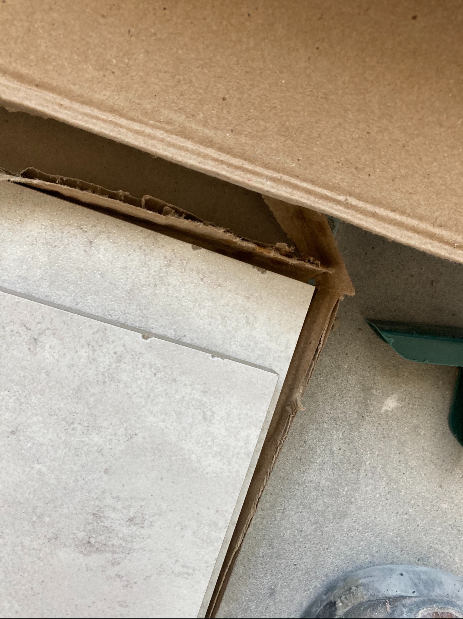 Paczka jasnych płytek ceramicznych w kartonowym opakowaniu, widoczny fragment zielonego narzędzia i buta na szarym podłożu.