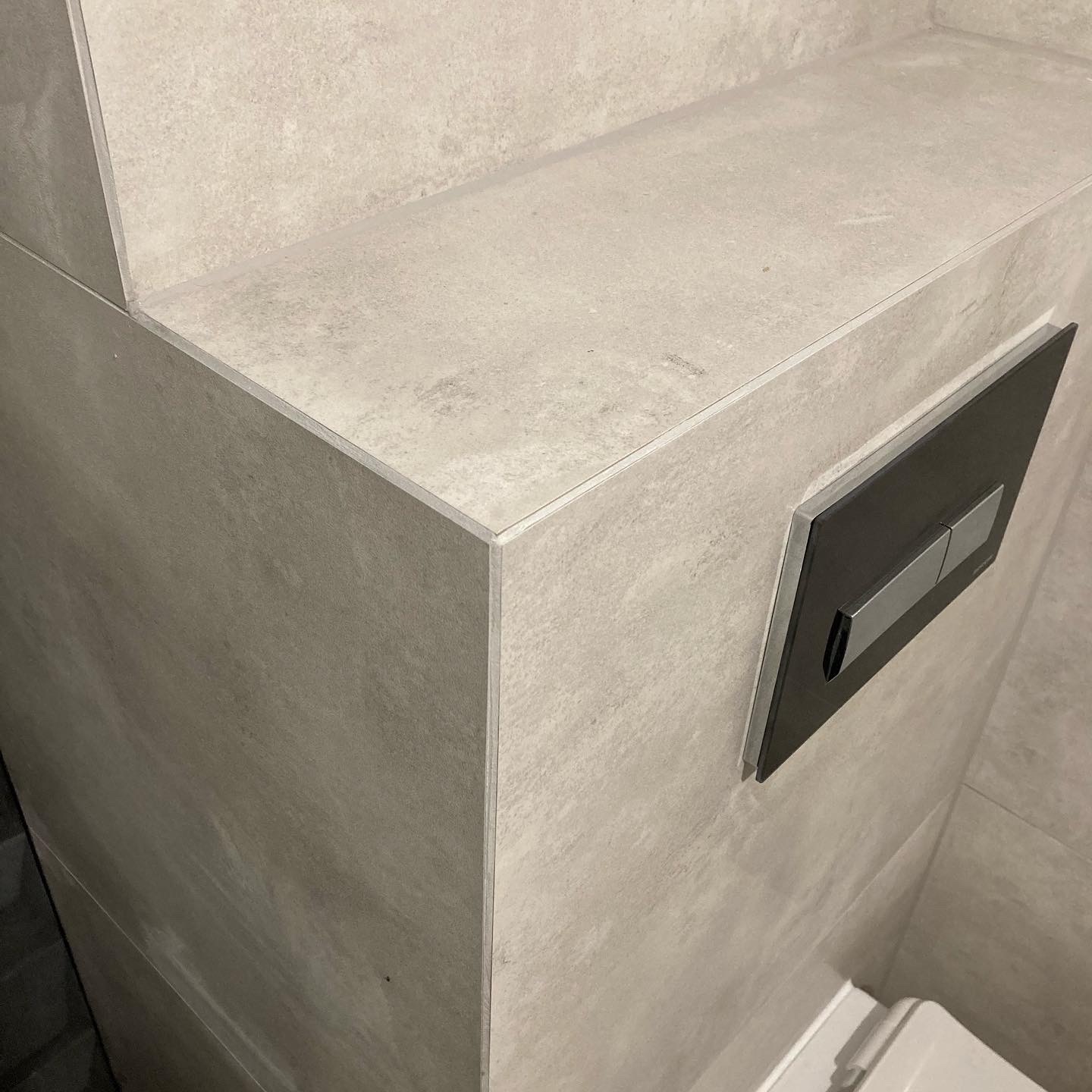 Wykończona ściana w łazience w Krakowie, pokryta dużymi szarymi płytkami ceramicznymi z widocznym przyciskiem spłuczki WC, z precyzyjnie wykonanymi narożnikami.