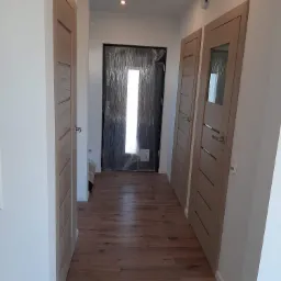 Korytarz z drewnianą podłogą, dwoje drzwi wewnętrznych z przeszkleniami i drzwi wejściowe zabezpieczone folią ochronną.