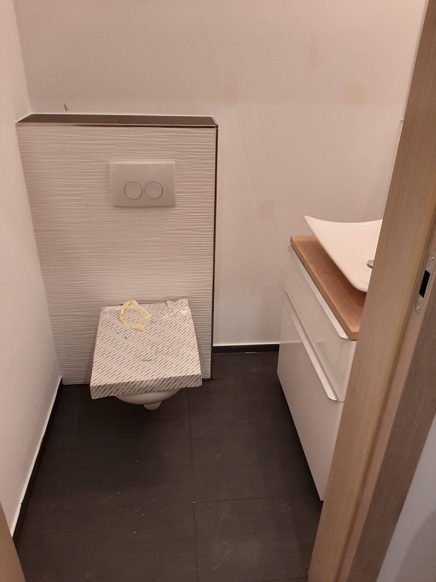 Nowoczesna łazienka w trakcie wykończenia: podwieszana toaleta z przyciskiem spłukującym na ścianie z dekoracyjną fakturą, umywalka nablatowa z drewnianym blatem, czarne płytki podłogowe.