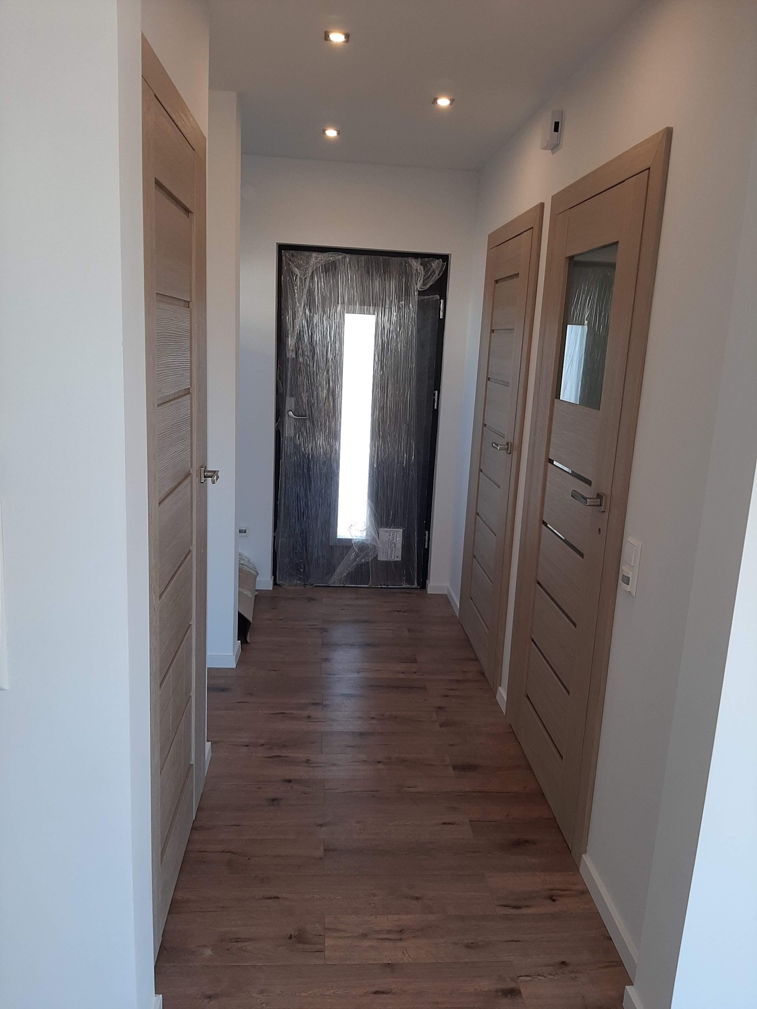 Korytarz z drewnianą podłogą, dwoje drzwi wewnętrznych z przeszkleniami i drzwi wejściowe zabezpieczone folią ochronną.