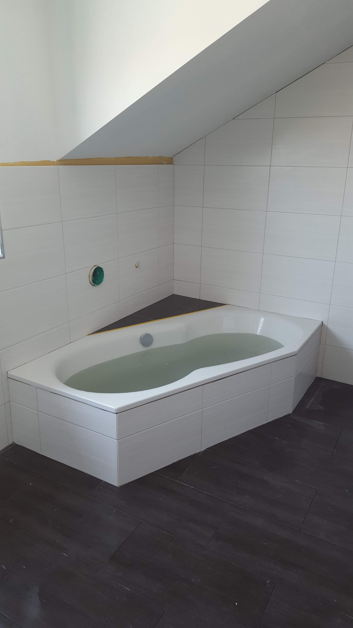 Wanna wypełniona wodą, obudowana białymi płytkami, w narożnej łazience na poddaszu z ciemną podłogą z płytek imitujących drewno.