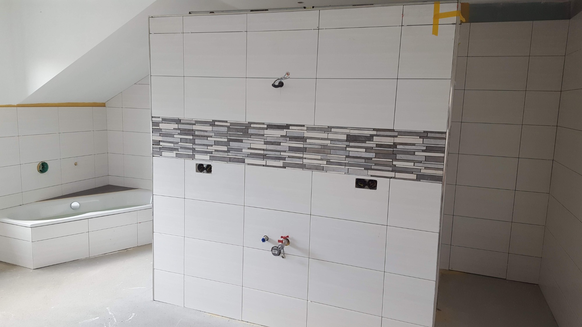 Wnętrze łazienki w trakcie wykańczania, białe płytki ścienne z dekoracyjnym pasem mozaiki, widoczne instalacje wodne i elektryczne, surowy beton na podłodze, wanna w rogu pomieszczenia.