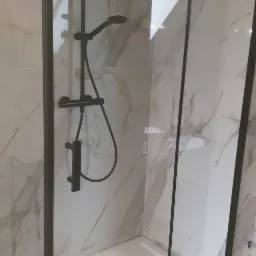 Kabina prysznicowa z czarnymi profilami i deszczownicą na tle marmurowych płytek, widoczny biały brodzik i butelka płynu do kąpieli