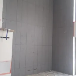 Montaż szarej okładziny ściennej z płyt w pionowym układzie, widoczne otwory na gniazdka elektryczne, fragment pomieszczenia w trakcie prac wykończeniowych.