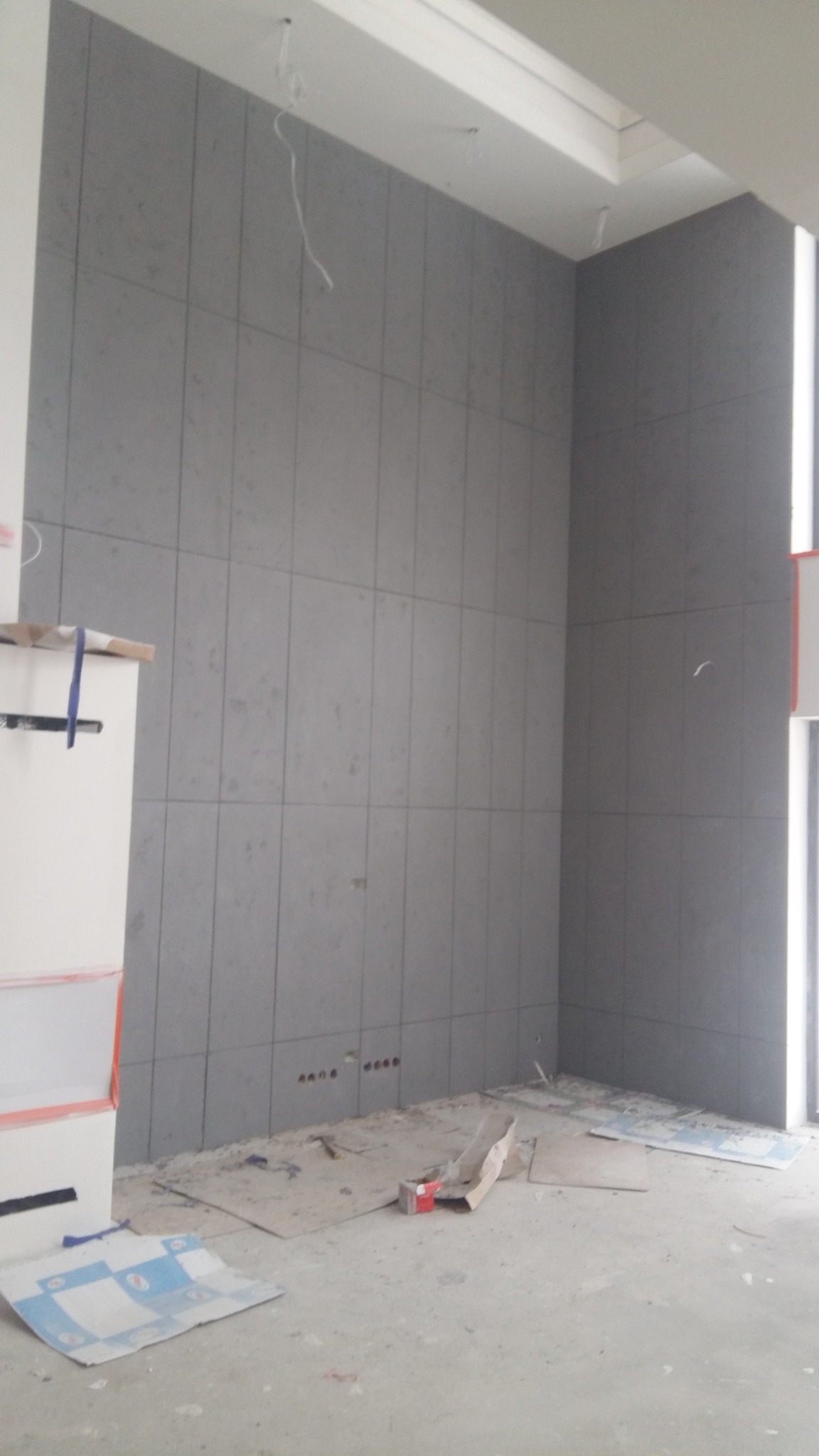Montaż szarej okładziny ściennej z płyt w pionowym układzie, widoczne otwory na gniazdka elektryczne, fragment pomieszczenia w trakcie prac wykończeniowych.