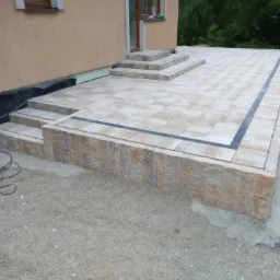 Jasna nawierzchnia z kostki brukowej, obrzeża z ciemnej kostki, schody prowadzące do drzwi budynku, widoczne ślady prac budowlanych, gruz i kable na ziemi.