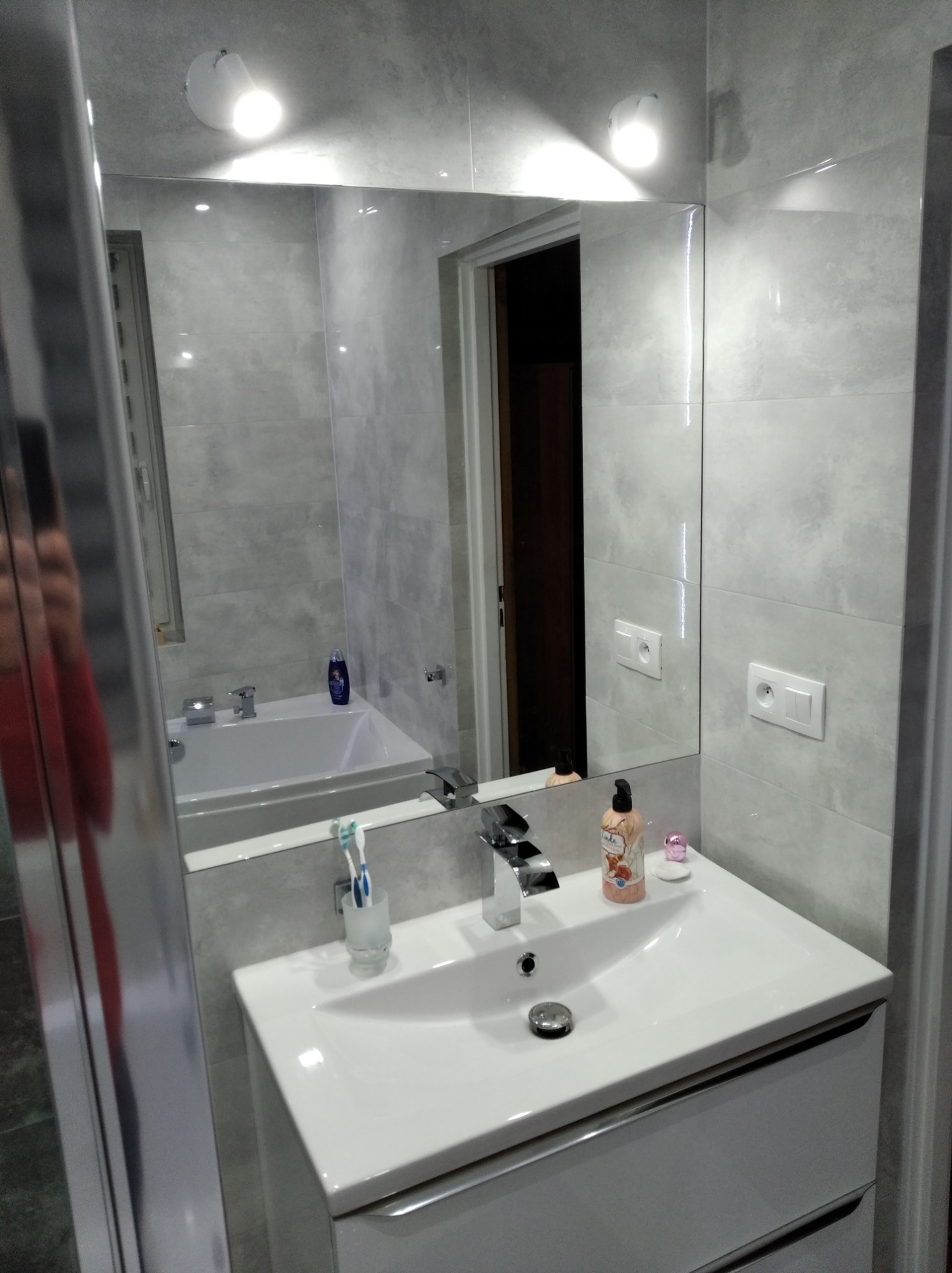 Umywalka z szafką w łazience wykończonej szarymi płytkami, widok w lustrze na wannę i drzwi