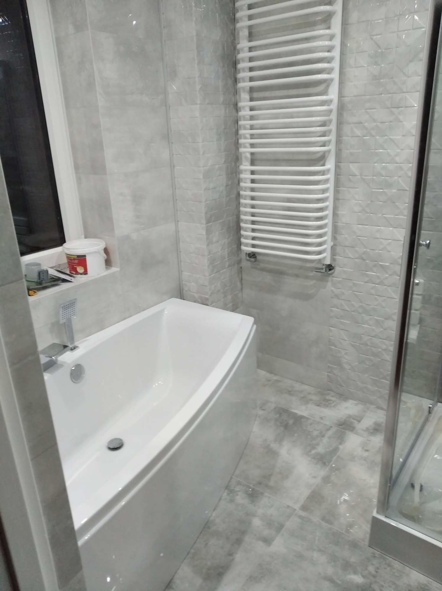 Nowoczesna łazienka w odcieniach szarości z białą wanną, grzejnikiem drabinkowym i kabiną prysznicową z widocznymi szarymi płytkami o wzorzystej fakturze.