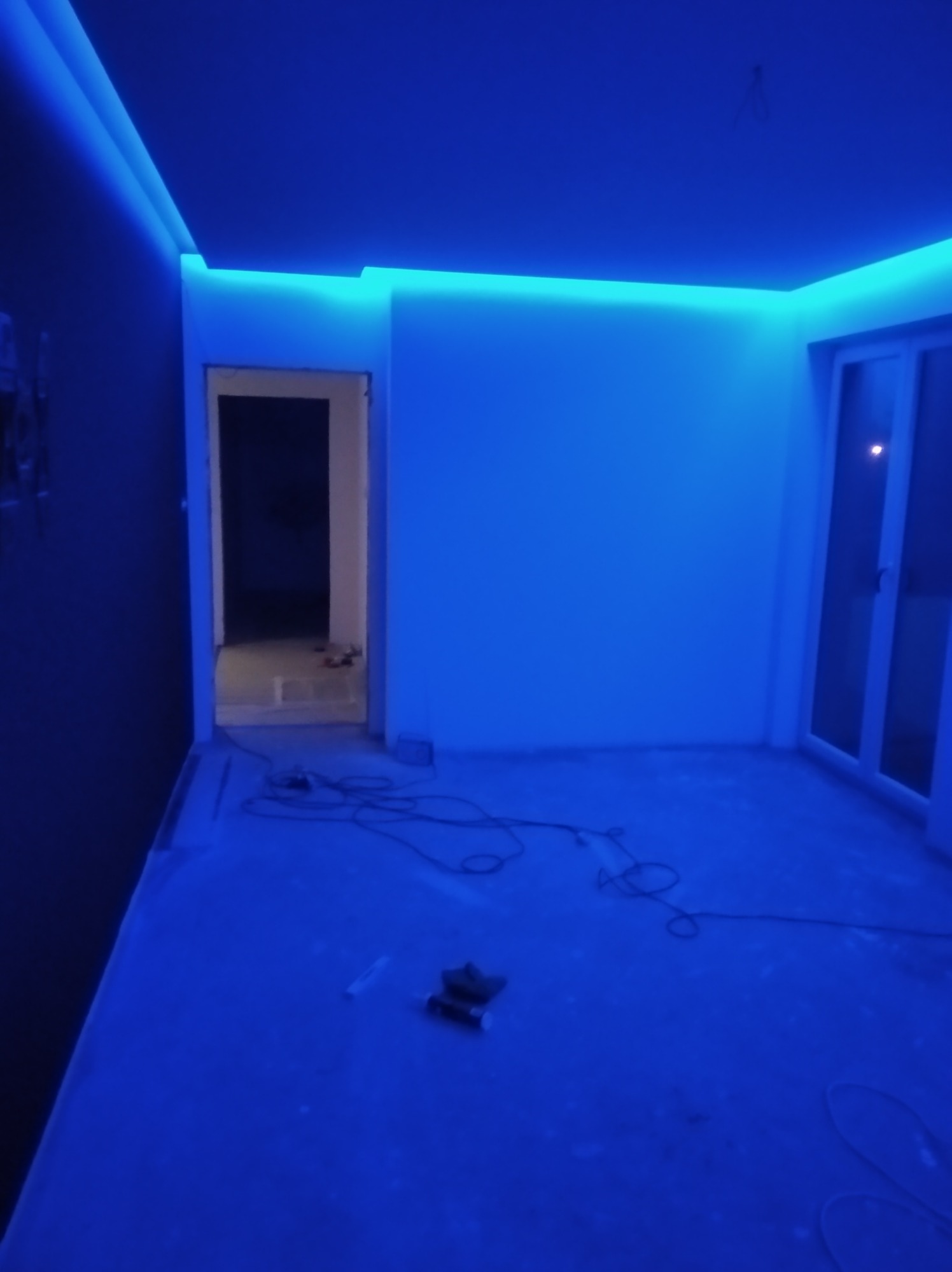 Pokój w trakcie remontu oświetlony intensywnym niebieskim światłem LED zamontowanym pod sufitem, widoczne narzędzia i kable na podłodze, otwarte drzwi do innego pomieszczenia.