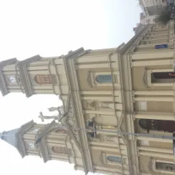 Fasada zabytkowego kościoła z dwiema wieżami zegarowymi, kolumnami, rzeźbami i zdobieniami, widziana z perspektywy ulicy z parkującymi samochodami.