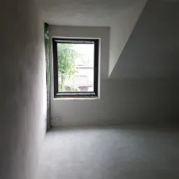 Wnętrze poddasza po nałożeniu szarego tynku maszynowego, widoczne okno z zielenią na zewnątrz oraz rura wentylacyjna przy ścianie.