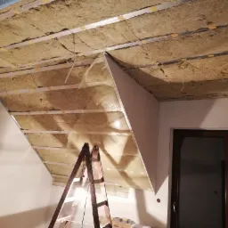 Ocieplenie poddasza wełną mineralną, widoczne metalowe profile, folia paroizolacyjna i częściowo zamontowana płyta kartonowo-gipsowa na skośnym suficie poddasza, drabina oparta o ścianę.