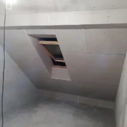 Poddasze z zamontowaną płytą gipsowo-kartonową, widoczne okno dachowe z drewnianą ramą i widoczne wkręty mocujące płyty.