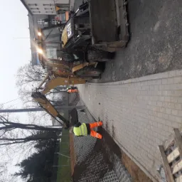 Żółta koparko-ładowarka CAT na placu budowy, pracownik w odblaskowej kamizelce przy układaniu kostki brukowej, widoczne elementy stabilizujące grunt. Ujęcie z góry, pochmurne niebo w tle.