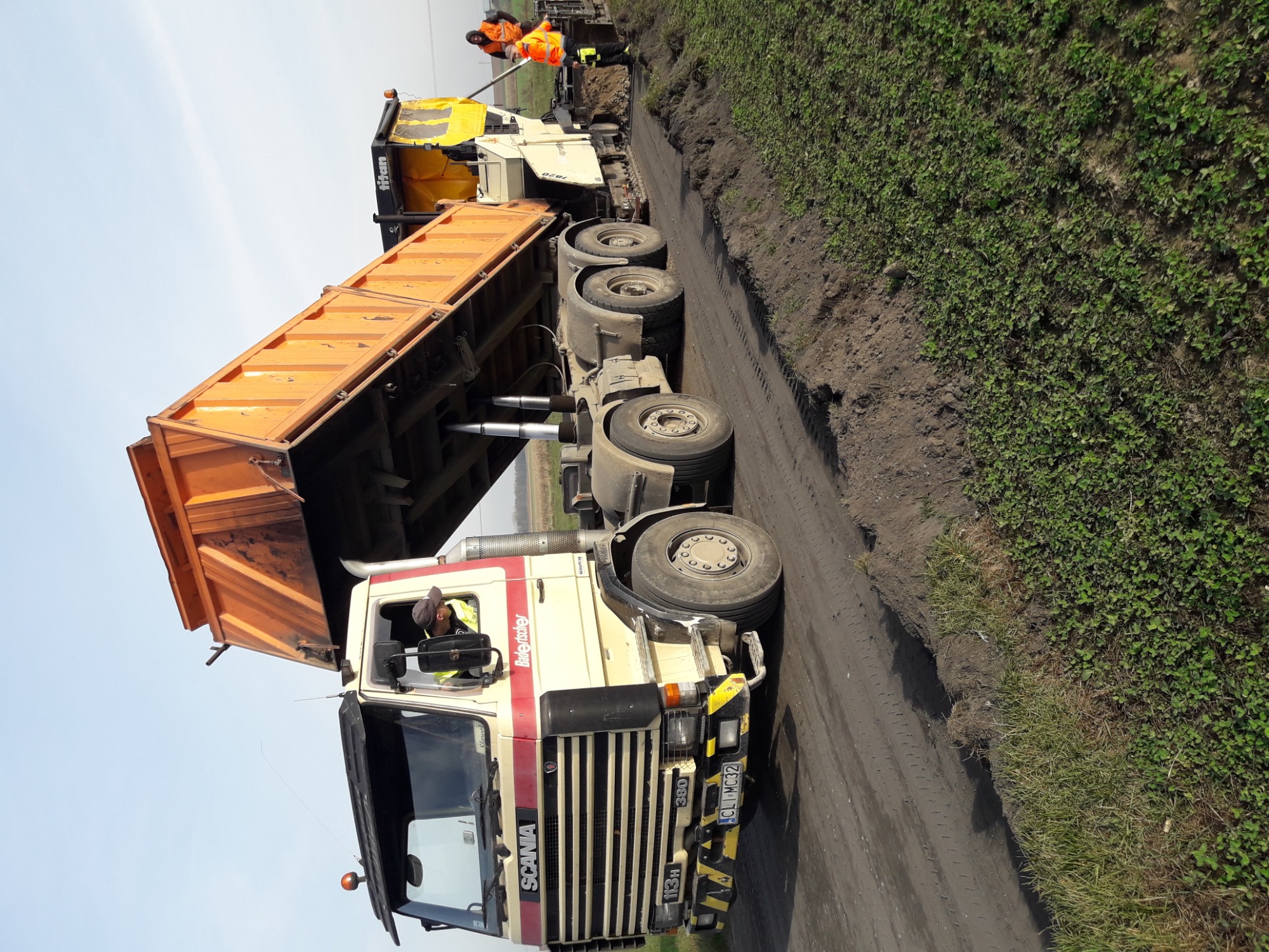 Pomarańczowa wywrotka Scania 113H z podniesioną skrzynią ładunkową, wysypująca materiał na nowo budowaną drogę, w tle maszyna do układania asfaltu i pracownik w pomarańczowym kombinezonie.