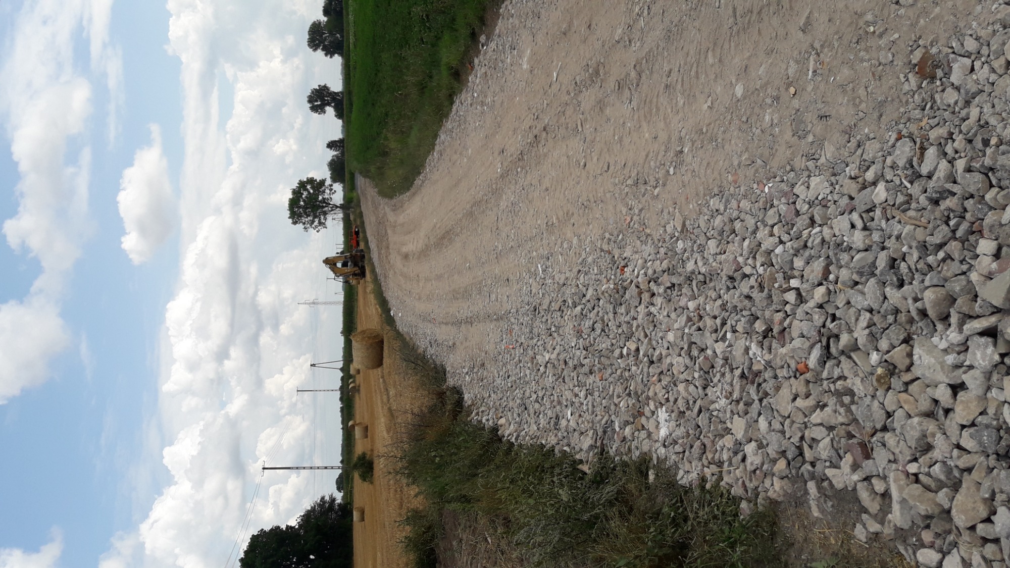 Nierówna, szutrowa droga polna w trakcie budowy lub naprawy, z widocznym fragmentem maszyny budowlanej w oddali, obok pole ze słomą w belach i słupami wysokiego napięcia, błękitne niebo z białymi...