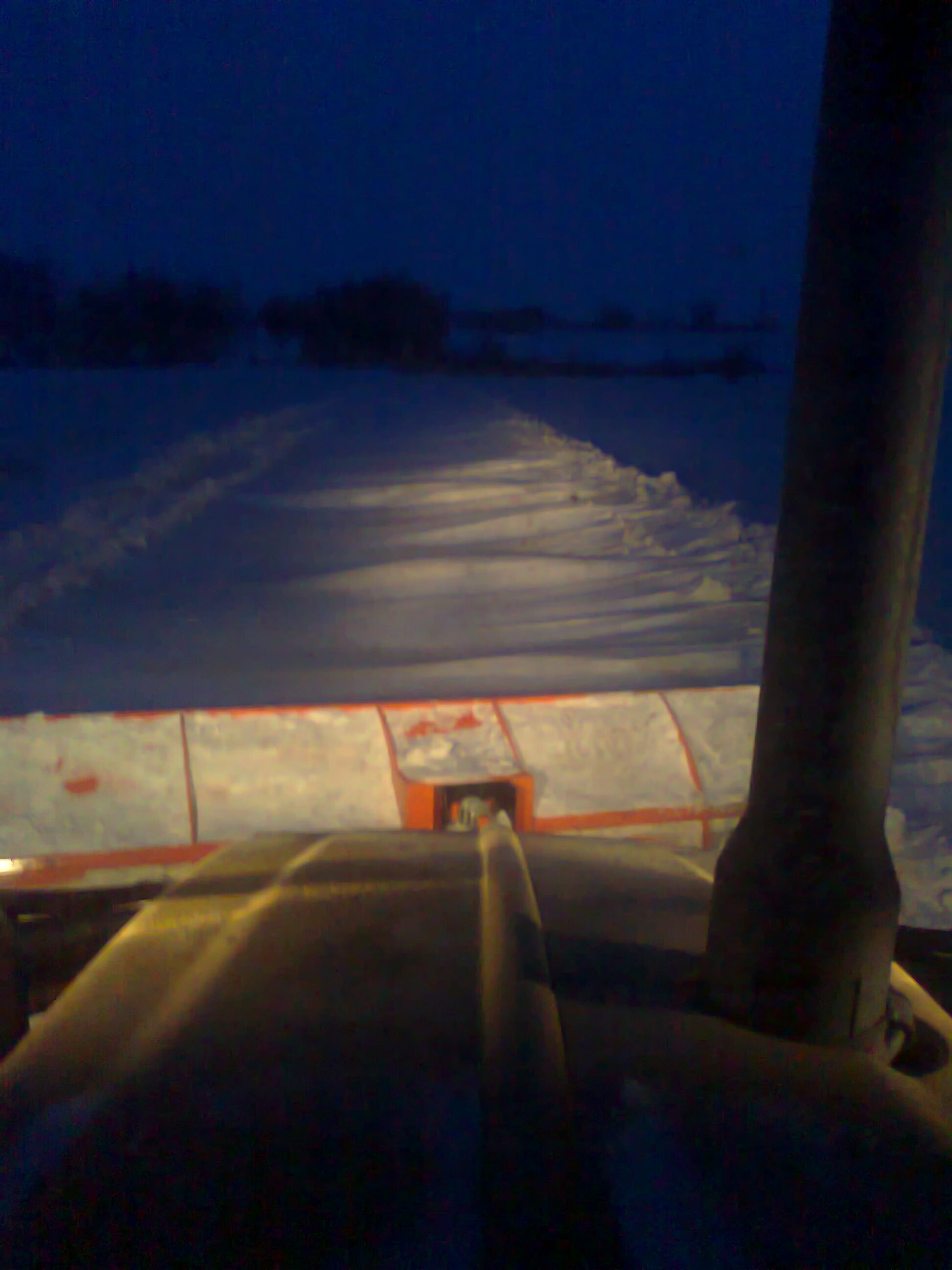 Widok z kabiny pojazdu na zaśnieżoną drogę, oświetloną reflektorami, z widocznym fragmentem pługu do odśnieżania.